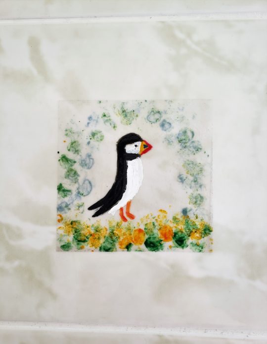 A puffin decal hand painted with a puffin and blue, green, yellow border. Photographed on a marble effect tile